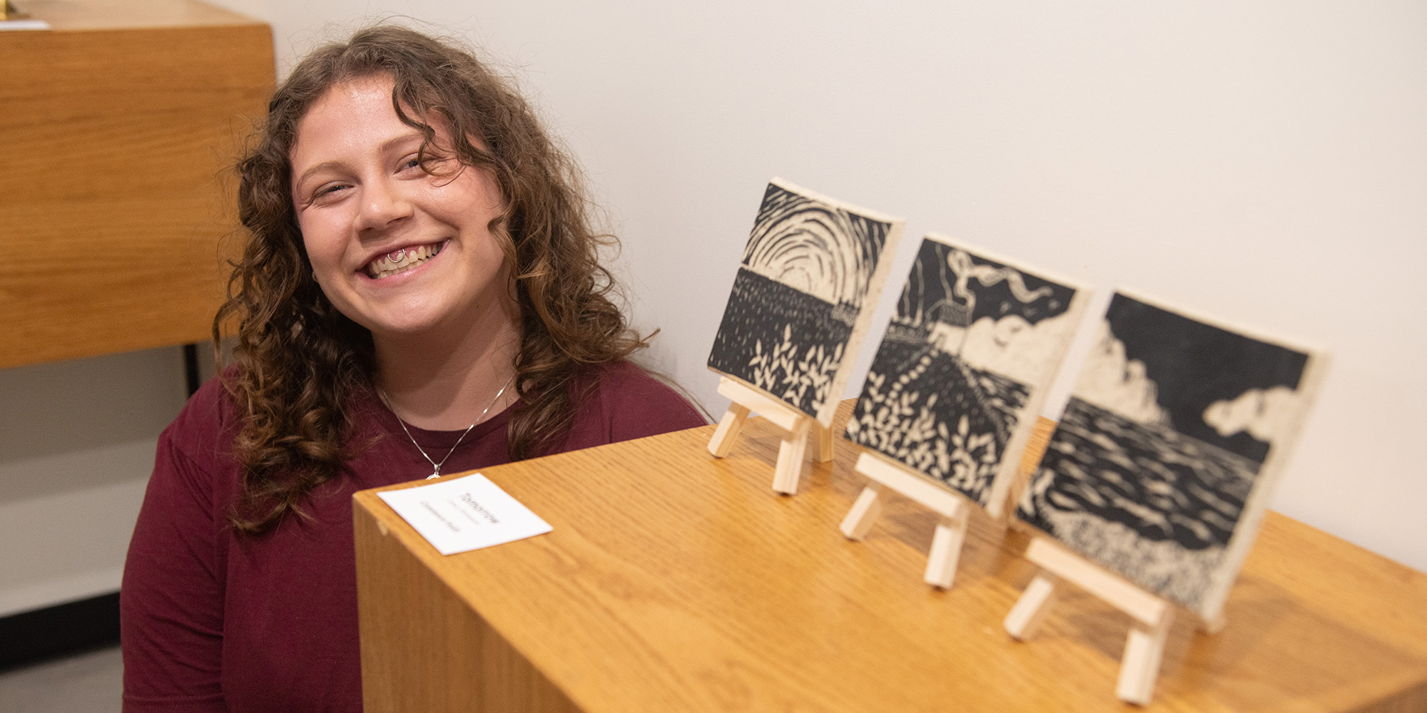 photo - Constance Feldt poses alongside her Best of Show-winning piece, “Tomorrow,” a ceramic tile triptych recognized at the 2026 Hesston College Annual Juried Student Art Exhibit. Stop by the Regier-Friesen Gallery through May 1 to see her work and the full exhibit on display.
