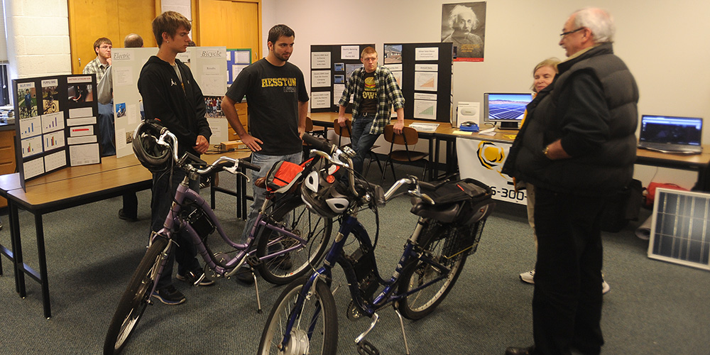 photo - Hesston College physics students Keenan Jensen, Hesston, Kan., and Ron Wenger, Adair, Okla., talk with Earth Day guests about the electric bicycles with solar power charging stations they helped build. Other students presented about a possible solar electric plan for the J.D. Charles Hall of Science and Arts and environmental biology and microbiology students presented about sustainability practices ranging from recycling to water conservation. Engineering students at Hesston have opportunities in their physics classes to create projects like the bikes and solar plans that give them hands-on experience for their future careers. The college also has an articulation agreement with Kansas State University-Salina for engineering technology students to transfer into and complete their bachelor’s degree in four total years of college.